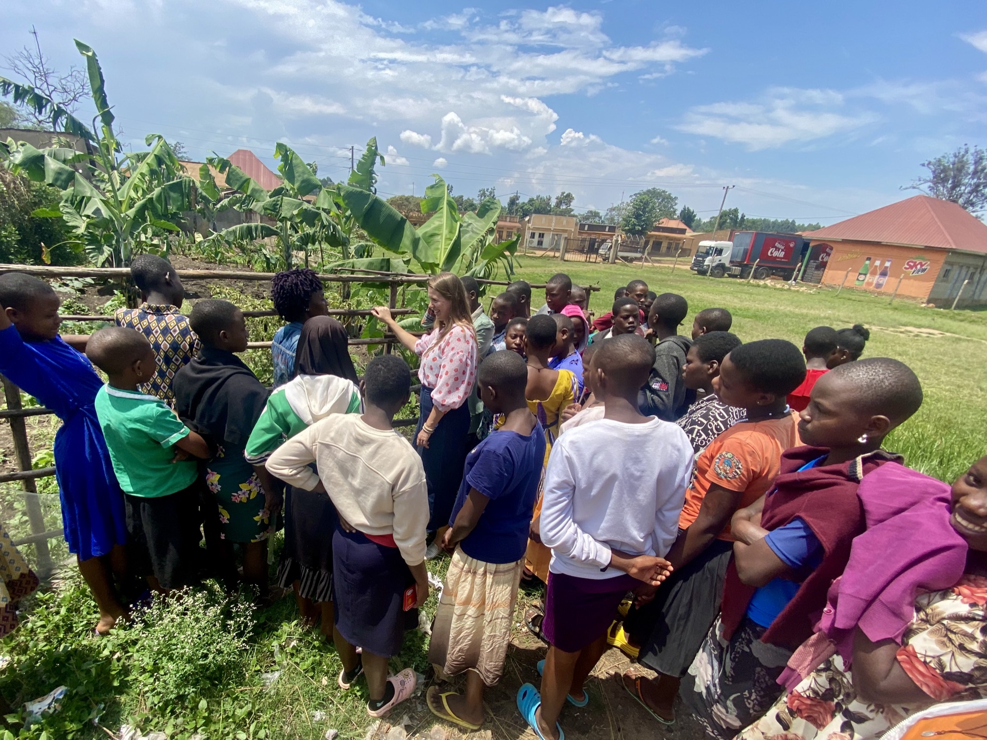 Community members and students gather at the fence of a food garden shaded by banana trees